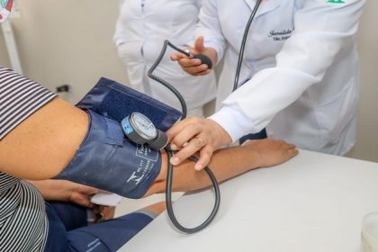 Imagem de braço com medidor de pressão arterial e profissional de saúde conferindo os dados do paciente.