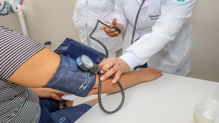 Imagem de braço com medidor de pressão arterial e profissional de saúde conferindo os dados do paciente. 